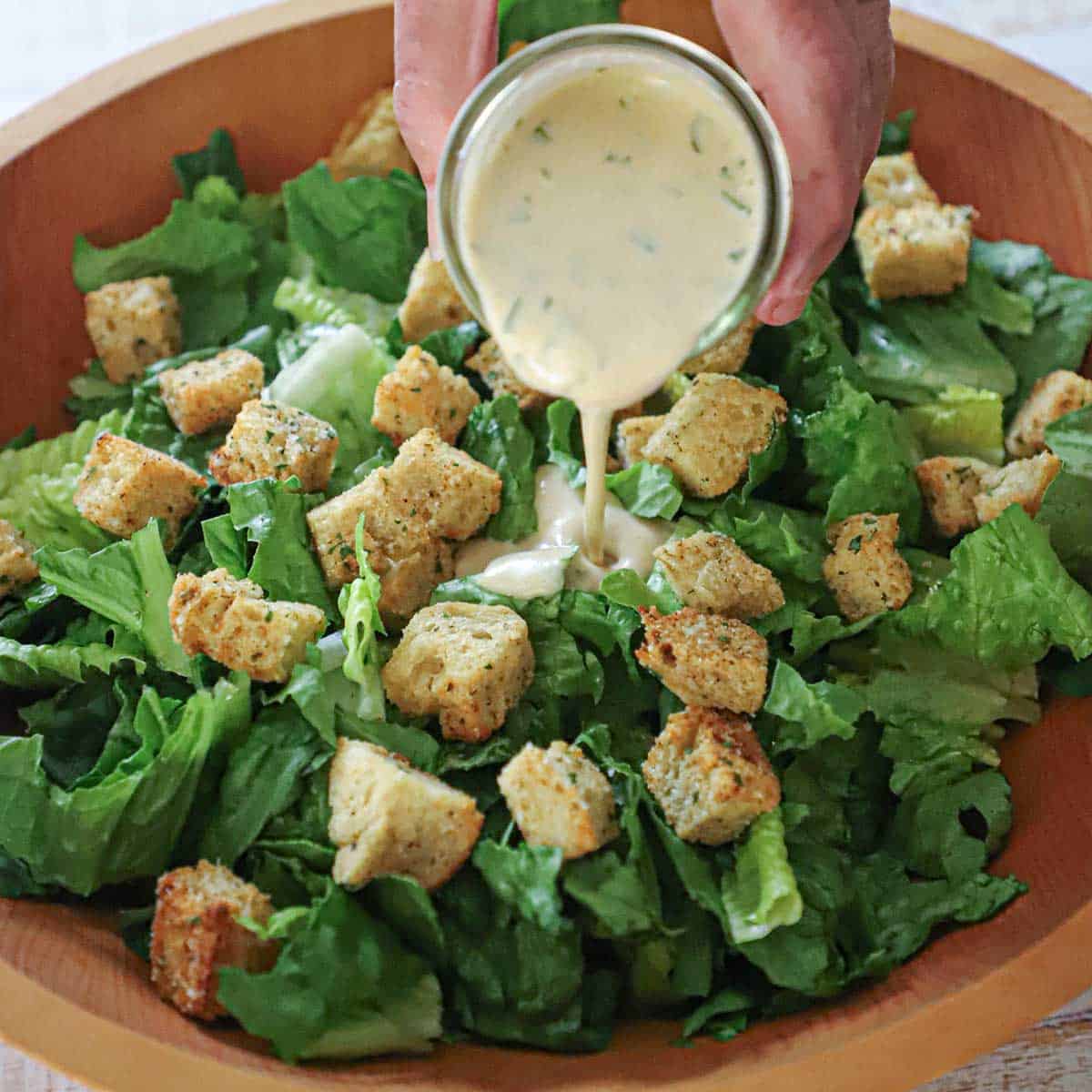 A person pouring homemade Caesar dressing over chopped romaine lettuce and homemade croutons in a large wooden salad bowl.