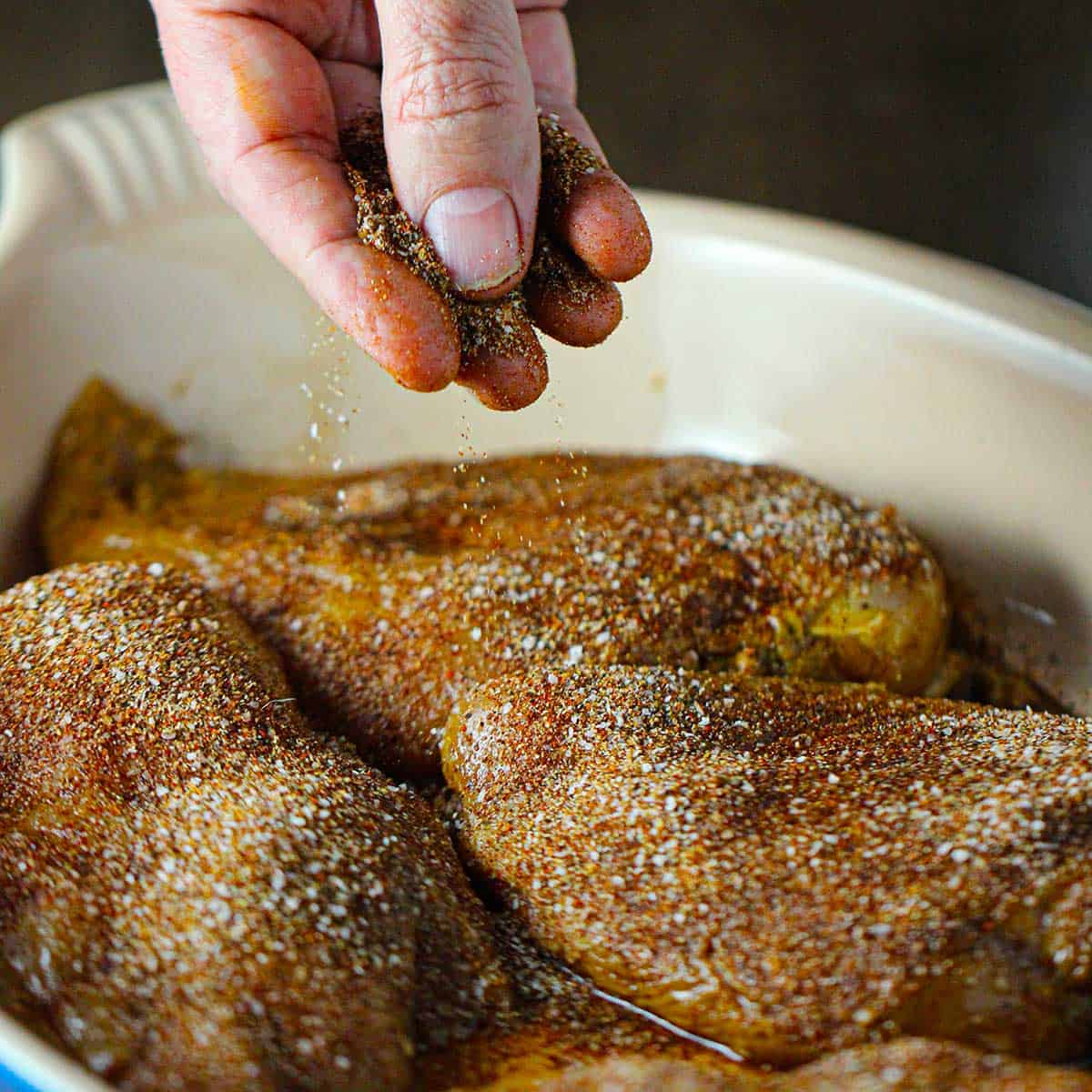 A person sprinkling fajita seasoning over skinless chicken breasts that have been marinated and are resting in an oval baking dish.