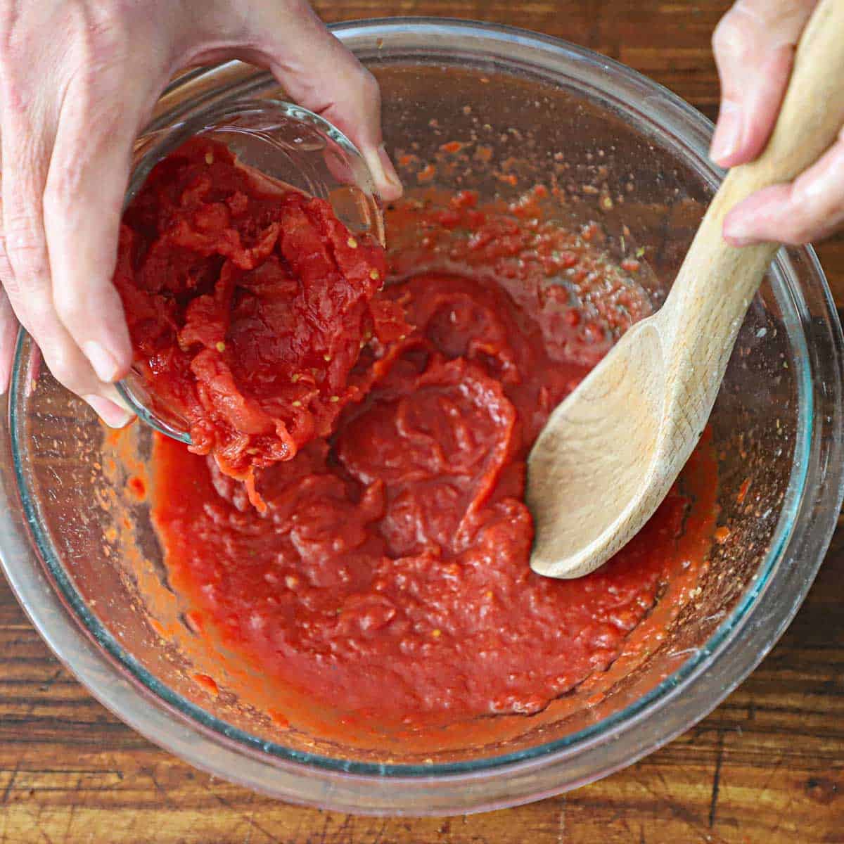 A person transferring hand-torn whole tomatoes into a bowl of ground tomatoes with a wooden spoon being held by his other hand.