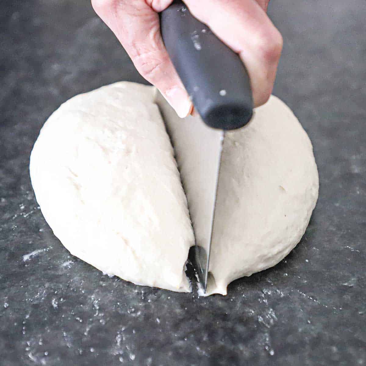 A person using a bench scraper to cut in half a ball of New York-style pizza dough on a black marble countertop.
