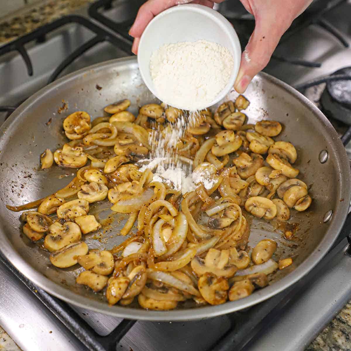 A person dumping all-purpose flour over sautéed onions and sliced mushrooms in a skillet on a gas stove.