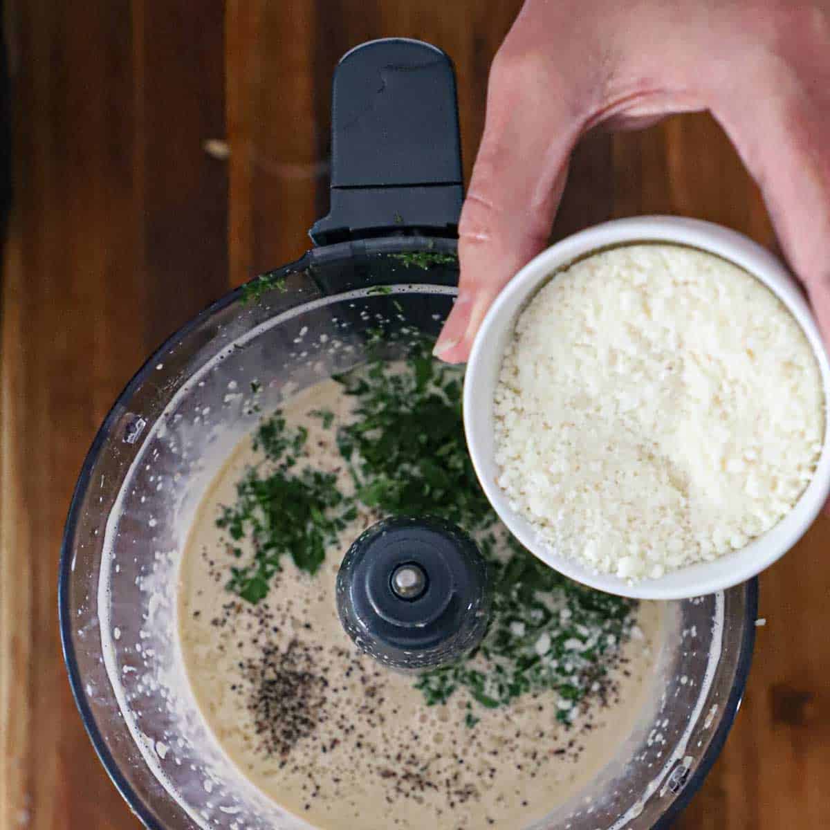 A person holding a small white bowl of grated Parmesan cheese over the bucket of a food processor that is filled with purée egg yolks, lemon juice, seasonings, and chopped Italian parsley.