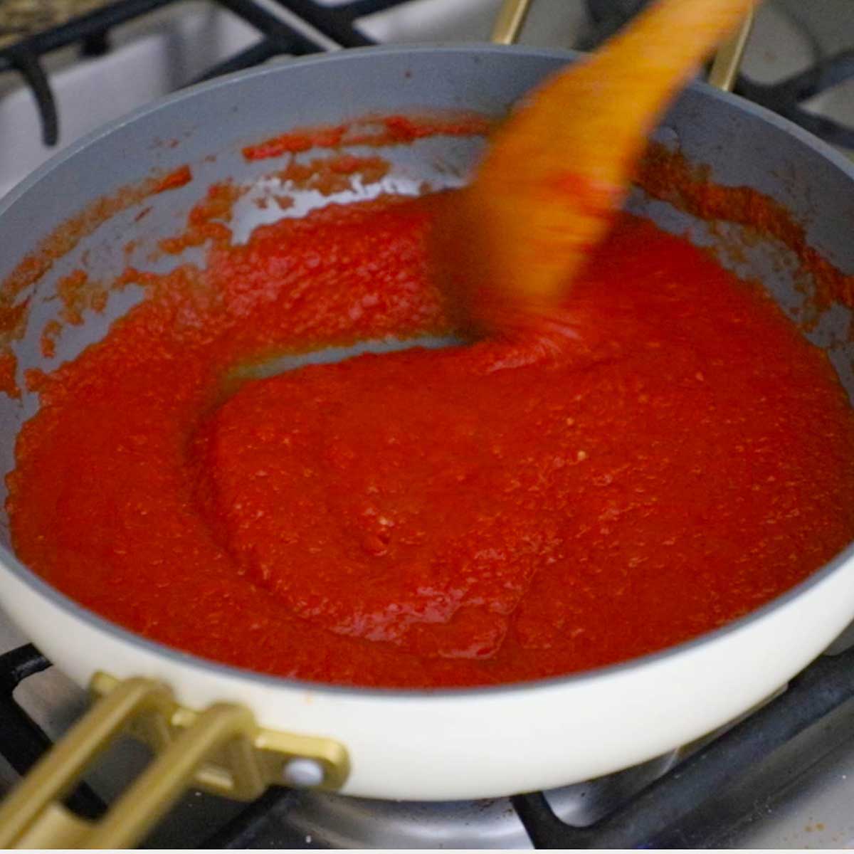 A person using a wooden spoon to stir simmering easy homemade ketchup in a large deep skillet on a gas stove.
