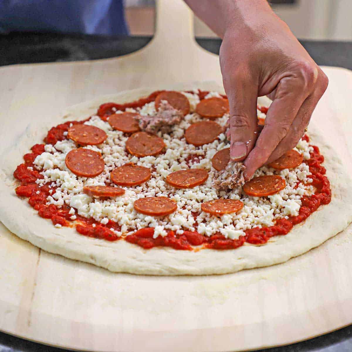 A person placing a small piece of uncooked ground pork onto an uncooked homemade New York-style pizza on a large pizza wheel.
