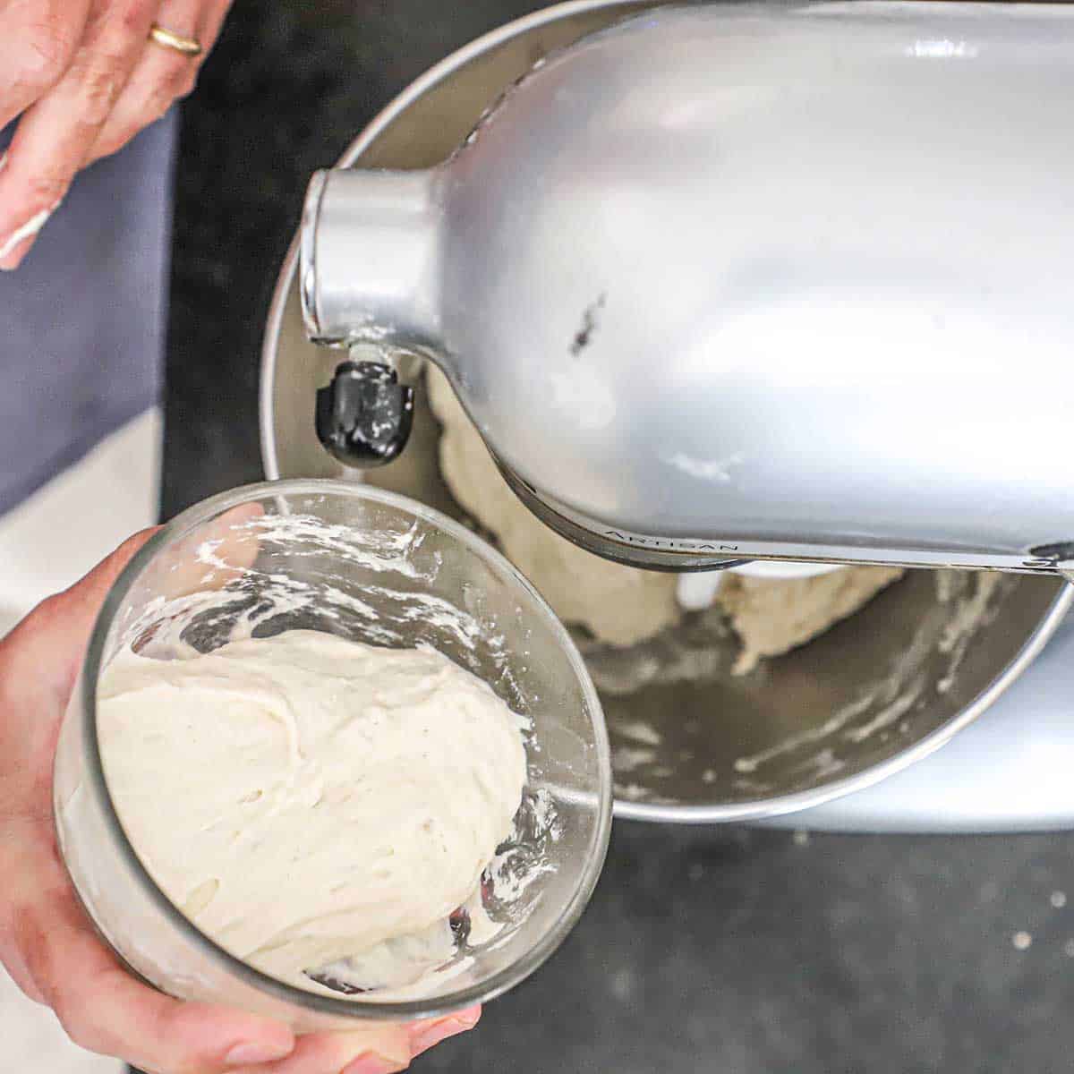 A person holding a small glass bowl filled with poolish, a pizza dough starter, over the bowl of a stand-mixer filled with the combined ingredients for New York-style pizza dough.
