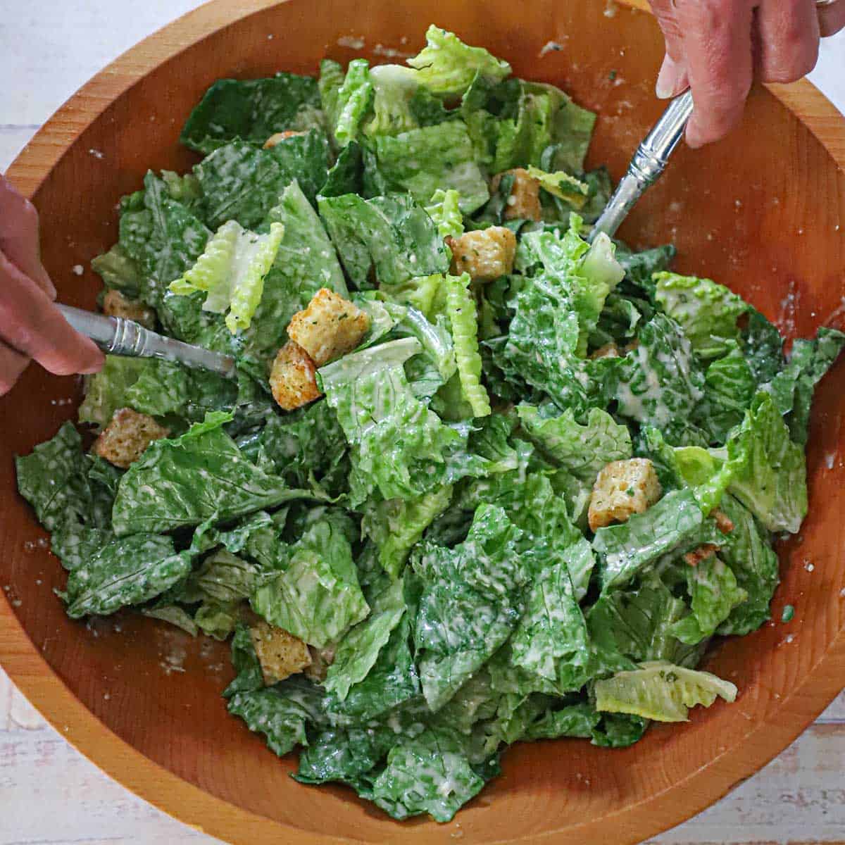 A person using two large silver serving spoons to toss together a homemade Caesar salad in a large wooden salad bowl.