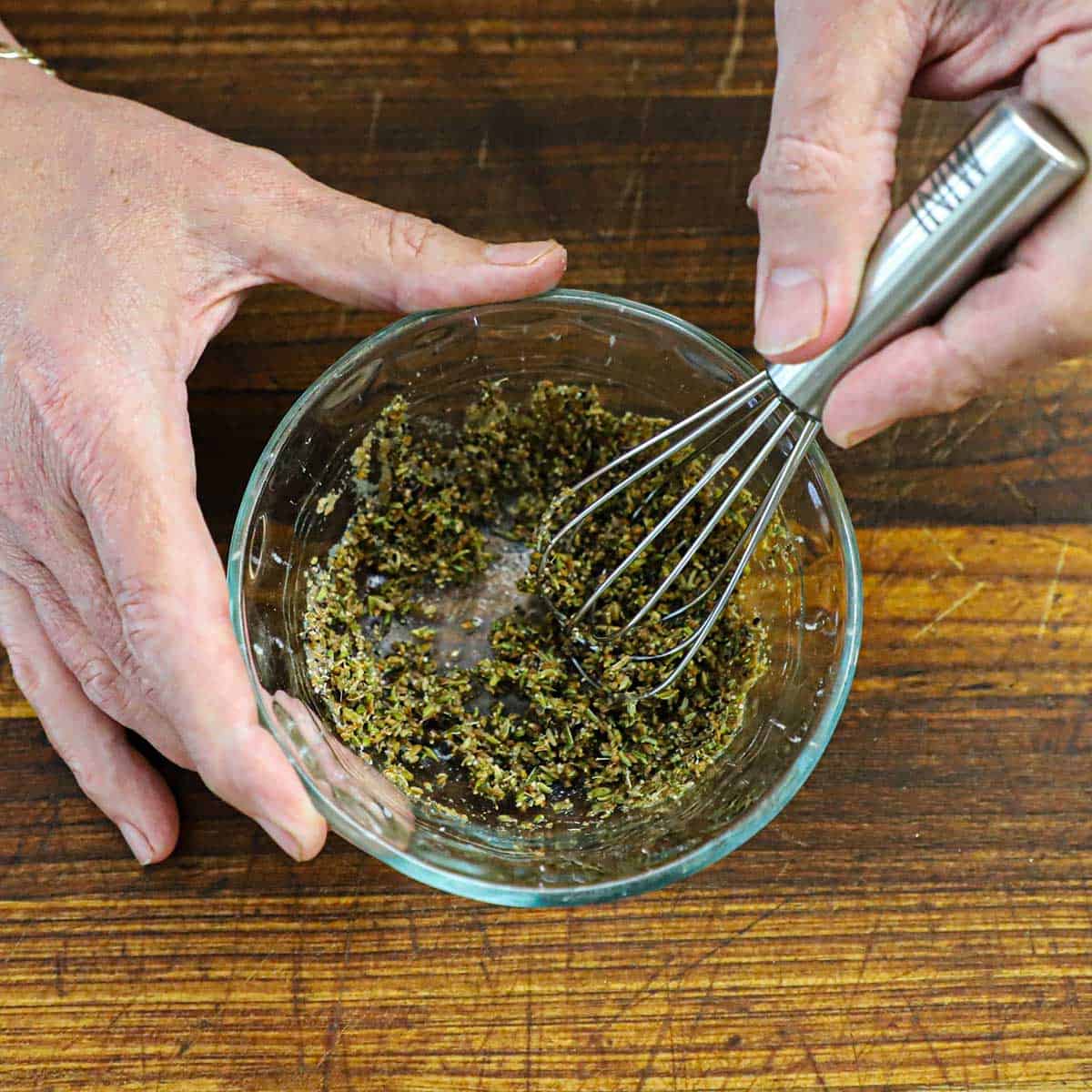 A person using a small whisk to incorporate water into a mixture of dried fennel, anise seeds, salt, and pepper in a small glass bowl.