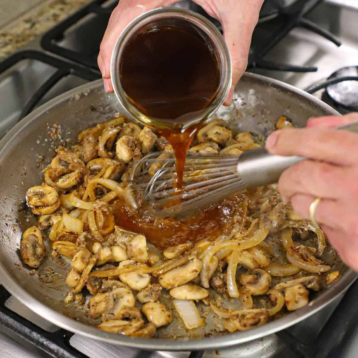 A person pouring beef broth from a glass jar into a skillet and is being whisked into sautéd onions and mushrooms that have been coated with flour.