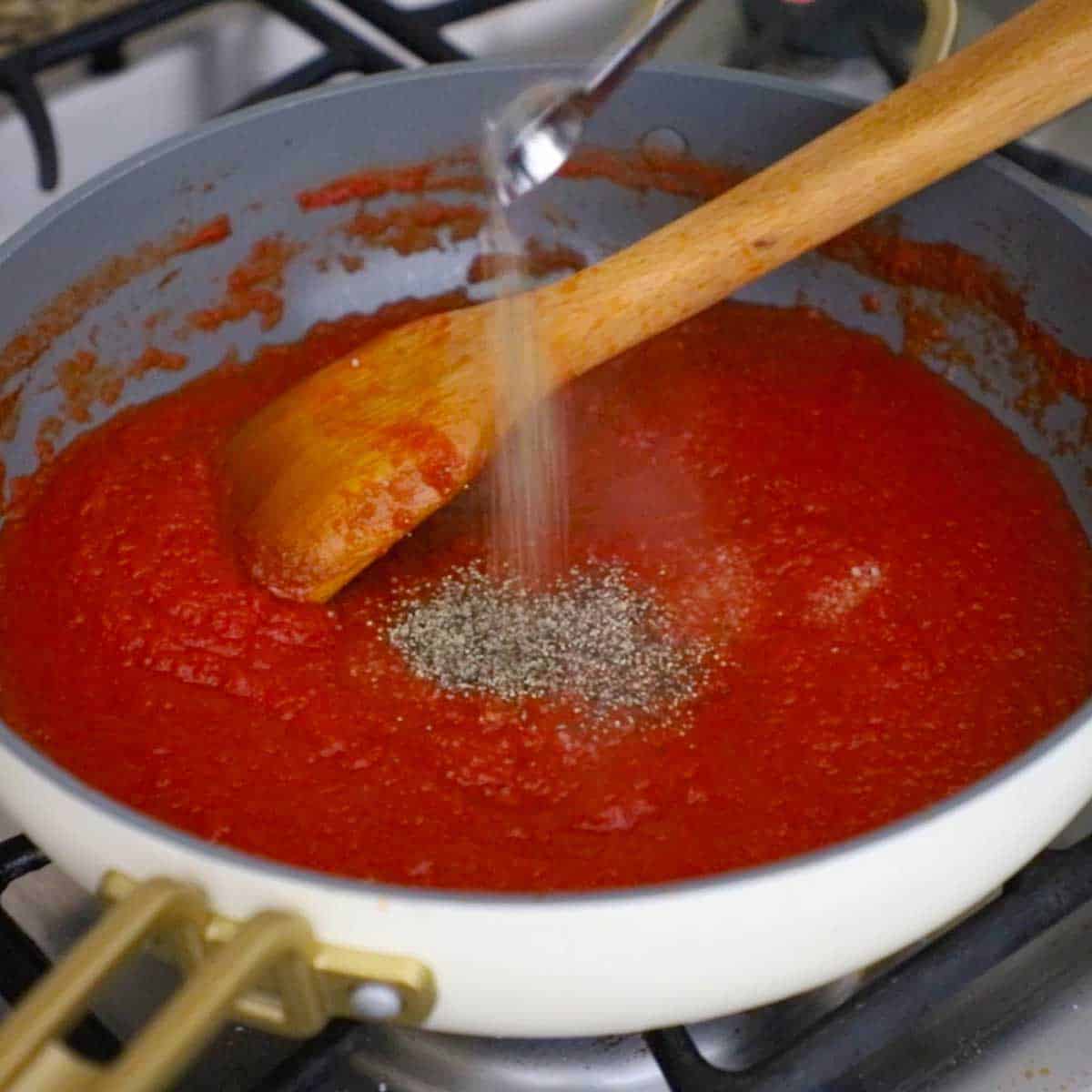 A person dumping ground black pepper from a teaspoon into a skillet filled with simmering easy homemade ketchup.