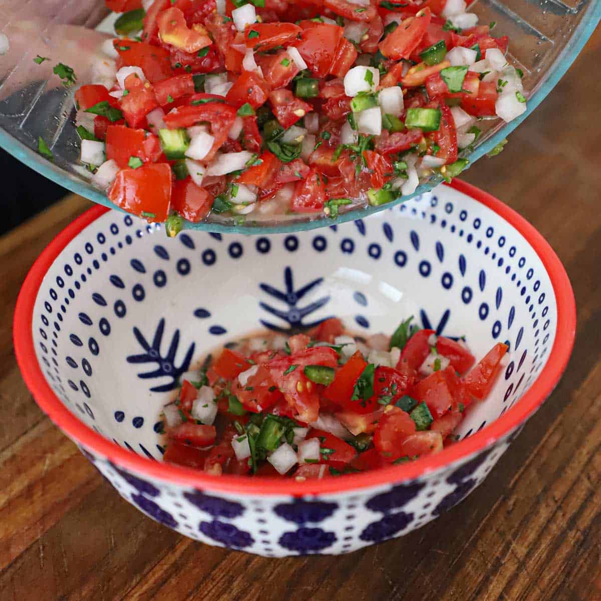 A person transferring homemade pico de gallo from a glass bowl into a colorful serving bowl sitting on a wooden cutting board.