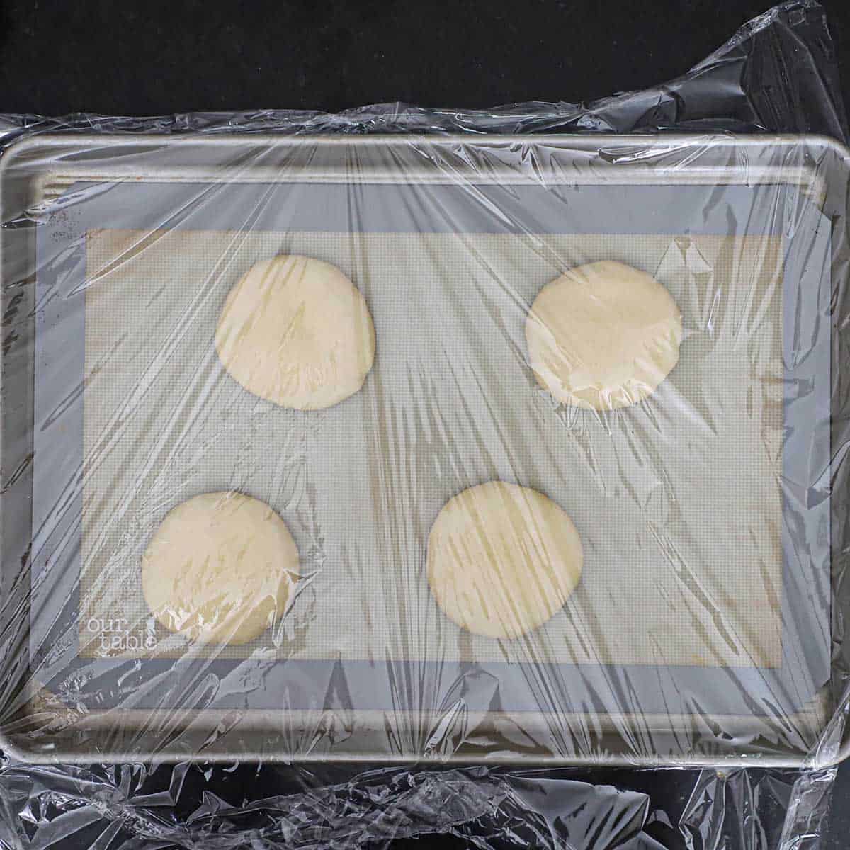 Four flattened rounds of hamburger bun dough on a silicone matt-lined sheet pan and is loosely covered with plastic wrap.