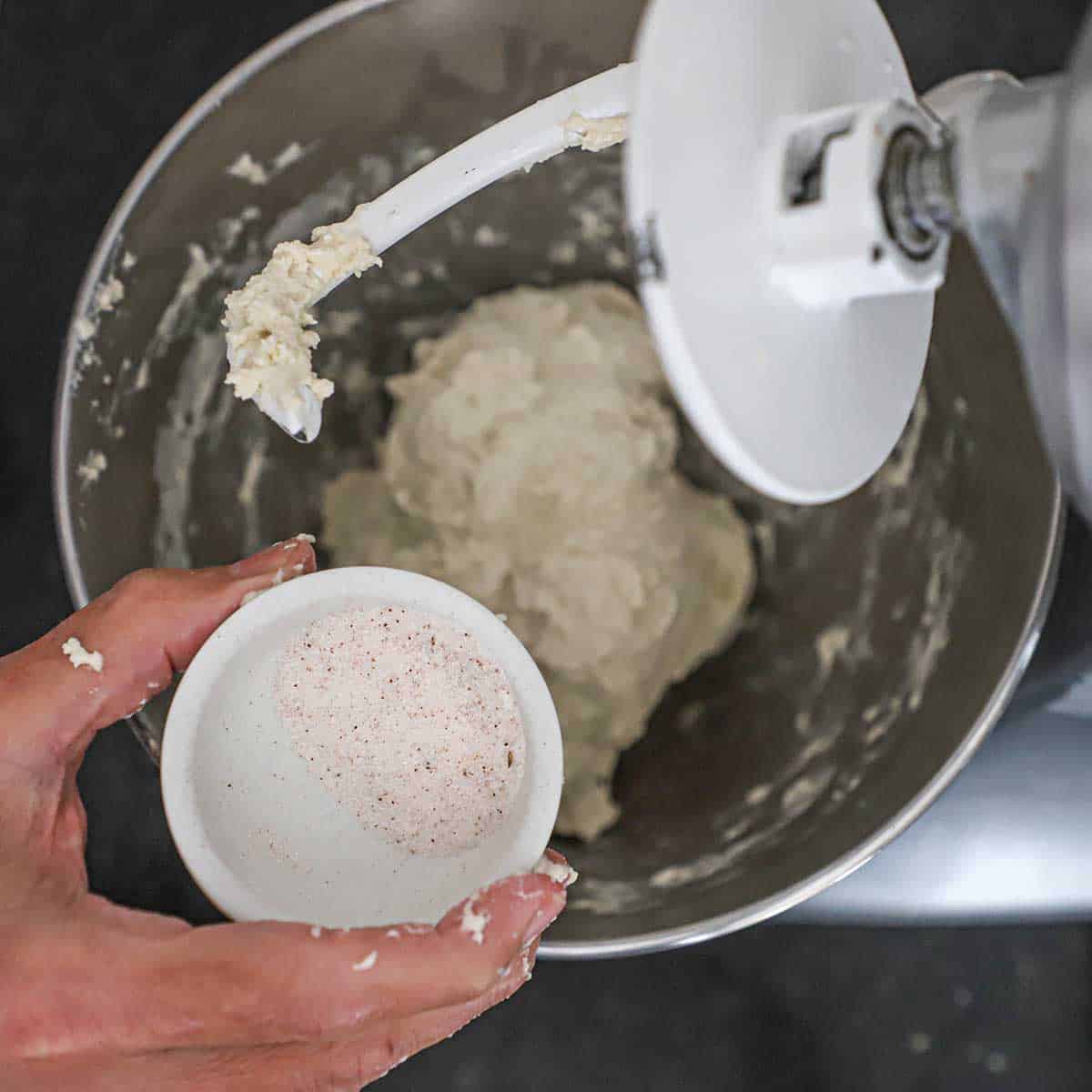 A person holding a small bowl of fine sea salt over the bowl of a stand-mixer filled with the ingredients of New York-style pizza dough.