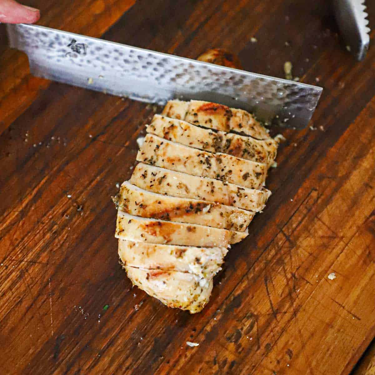 A person using a large Japanese knife to slice a grilled boneless, skinless chicken breast on a wooden cutting board.