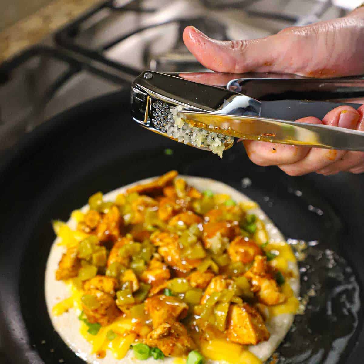 A person pressing garlic through a garlic press over the inside of a chicken quesadilla being cooked in a skillet with hot oil.