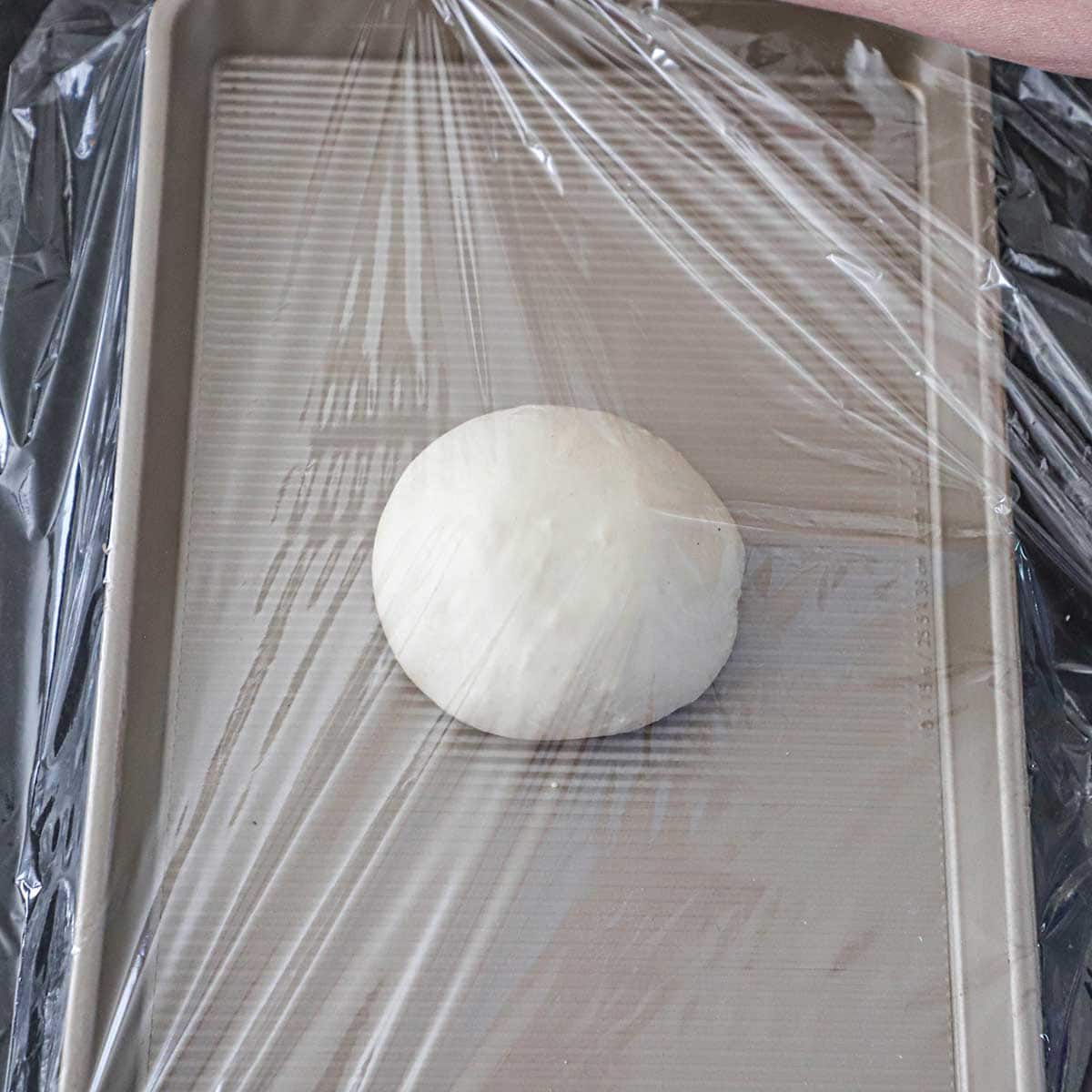 A smooth ball of New York-style pizza dough on a gold sheet pan wrapped in clear plastic wrap.
