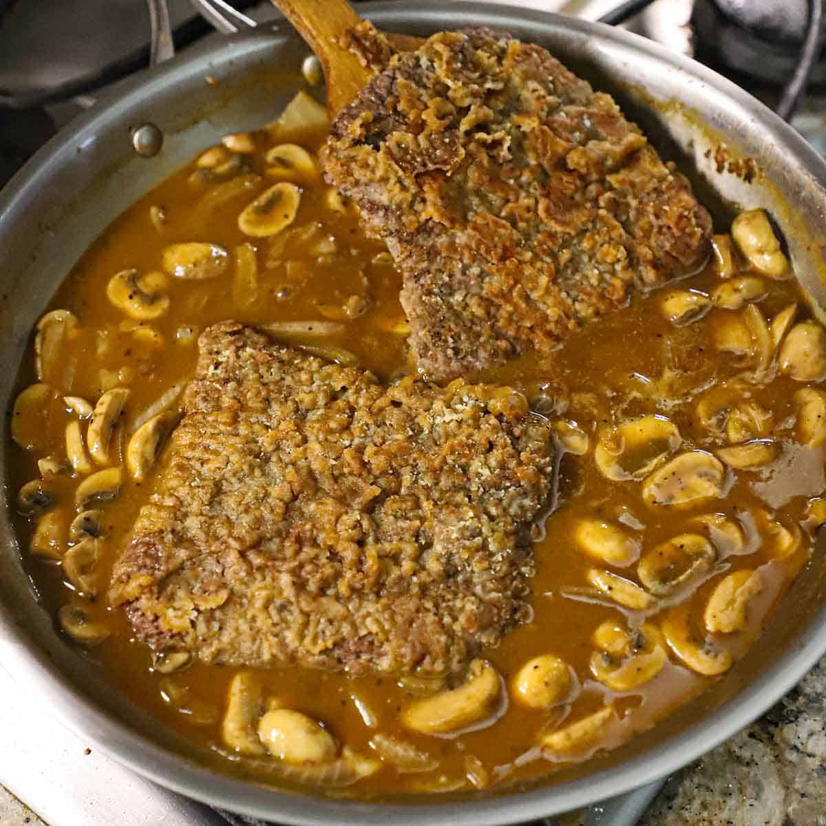 A person using a wooden spatula to place a browned cube steak into a skillet filled with a brown mushroom gravy.