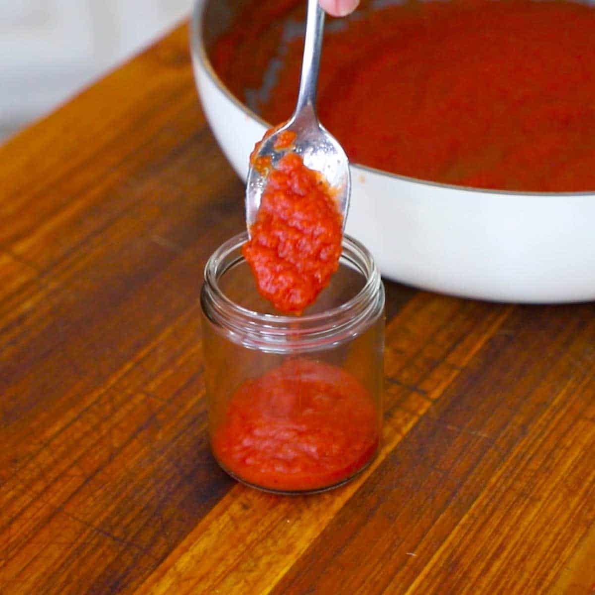 A person using a small spoon to transfer easy homemade ketchup into a small glass jar on a wooden cutting board.