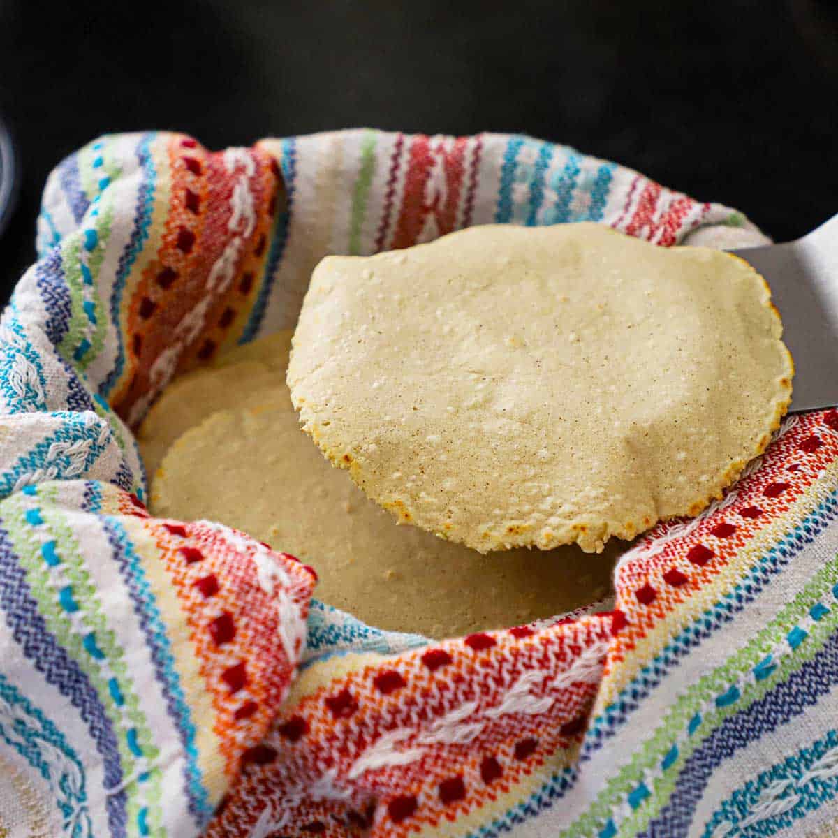 A person using a large metal spatula to place a freshly cooked corn tortilla into a tortilla warmer lined with a colorful napkin and is filled with other cooked tortillas.