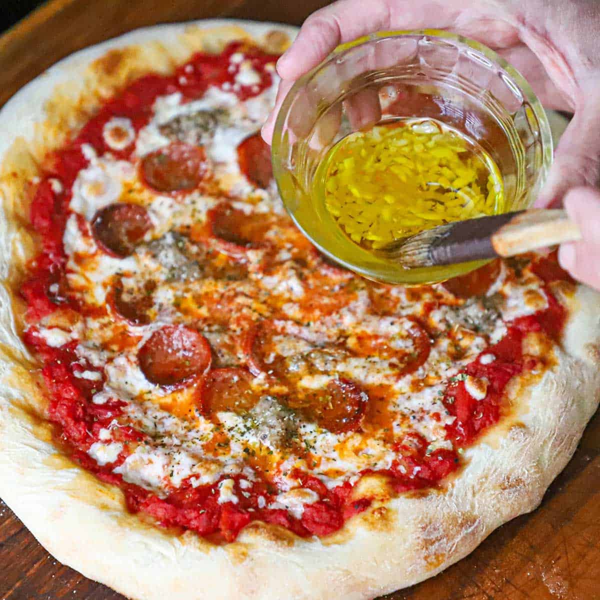 A person holding a brush that is in a bowl of garlic-infused olive oil over a freshly baked homemade New York-style pizza.