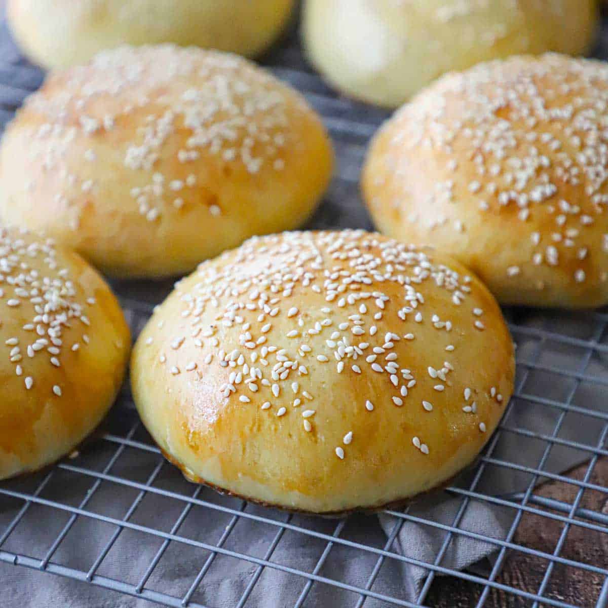 Several freshly baked homemade hamburger buns topped with sesame seeds resting on a baking rack over a grey cloth.