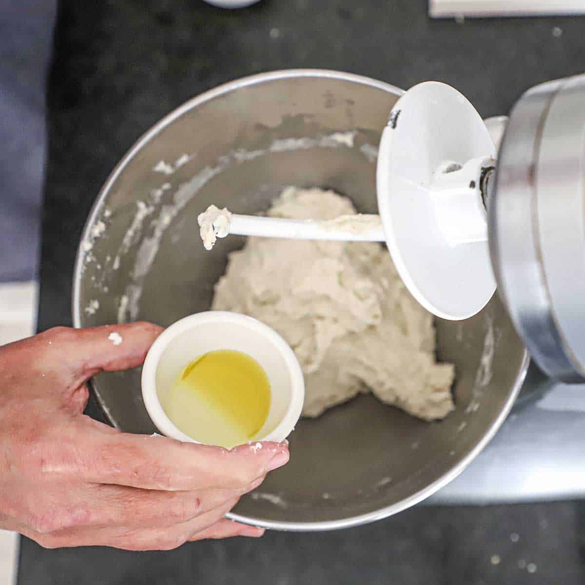A person holding a small bowl with a small amount of extra-virgin olive oil over a bowl of a stand-mixer filled with a New York-style pizza dough.