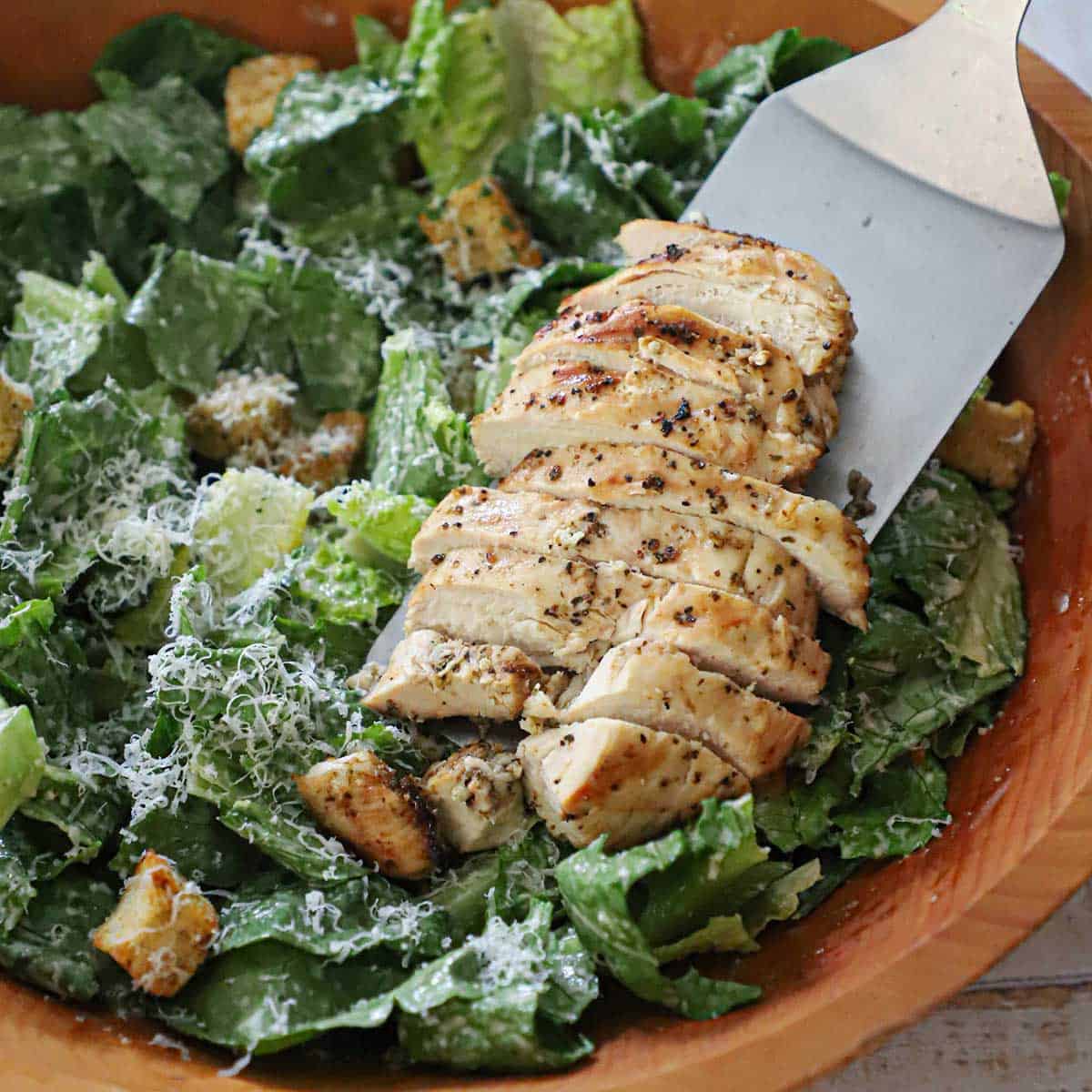 A person using a large silver spatula to place a sliced grilled chicken breast over a Caesar salad with homemade croutons in a large wooden salad bowl.