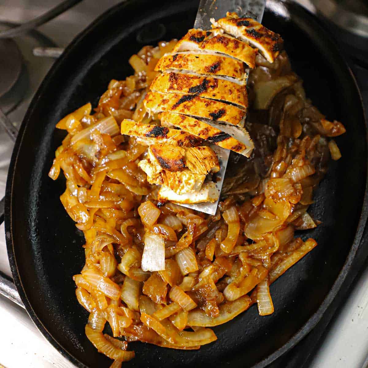 A person using a metal spatula to slide grilled sliced chicken onto a fajita pan filled with sizzling caramelized onions resting on a gas stovetop.