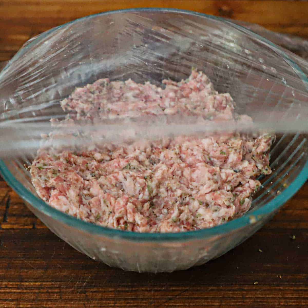 A person placing plastic wrap over a bowl of sweet feen sausage on a wooden cutting board.