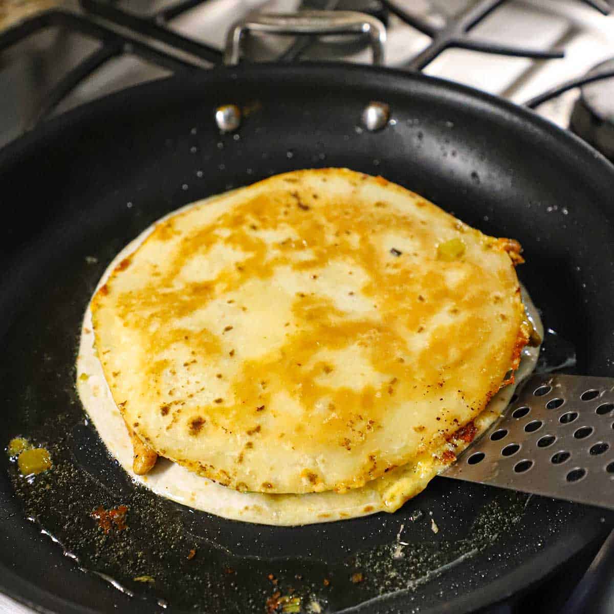 A person using a metal spatula to flip over a crispy chicken quesadilla that is simmering in oil in a large black skillet.