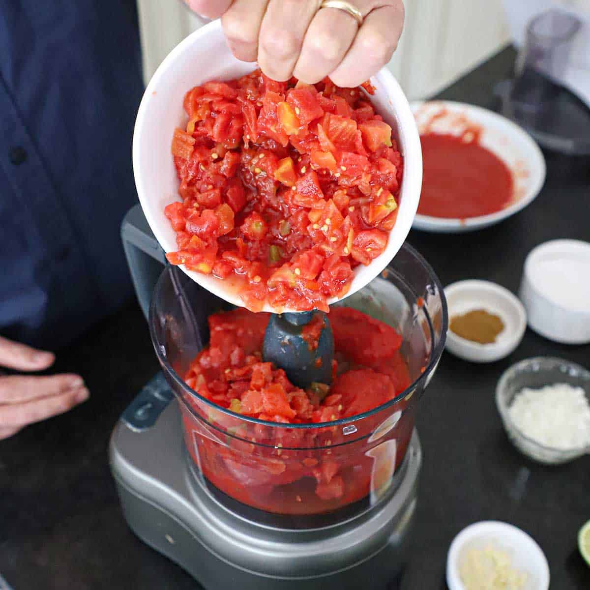 A person transferring diced tomatoes and chilis, also called Ro-tel, from a white bowl into the bucket of a food processor that is filled with whole tomatoes that were canned.