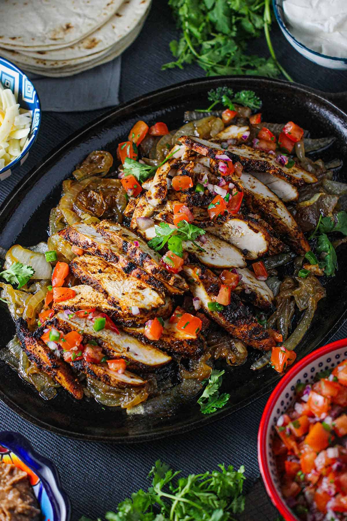 Tex-Mex chicken fajitas sitting on large oval fajita pan and topped with fresh pico de gallo and chopped cilantro with bowls of grated cheese and pico de gallo nearby.