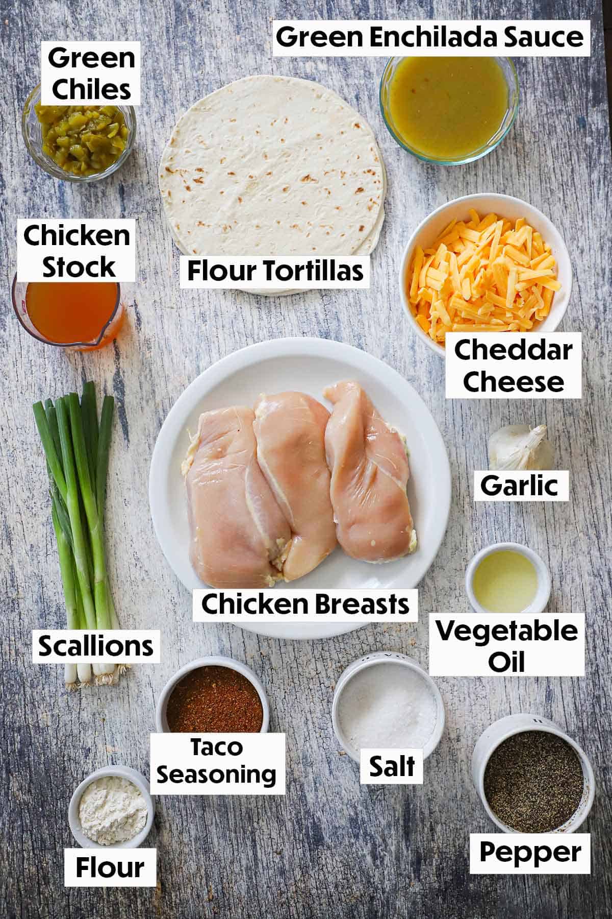 An arrangement of ingredients for chicken quesadilla on a grey wooden background including raw chicken breasts, tortillas, cheese, chicken broth, enchilada sauce, garlic, oil, flour, and seasonings.