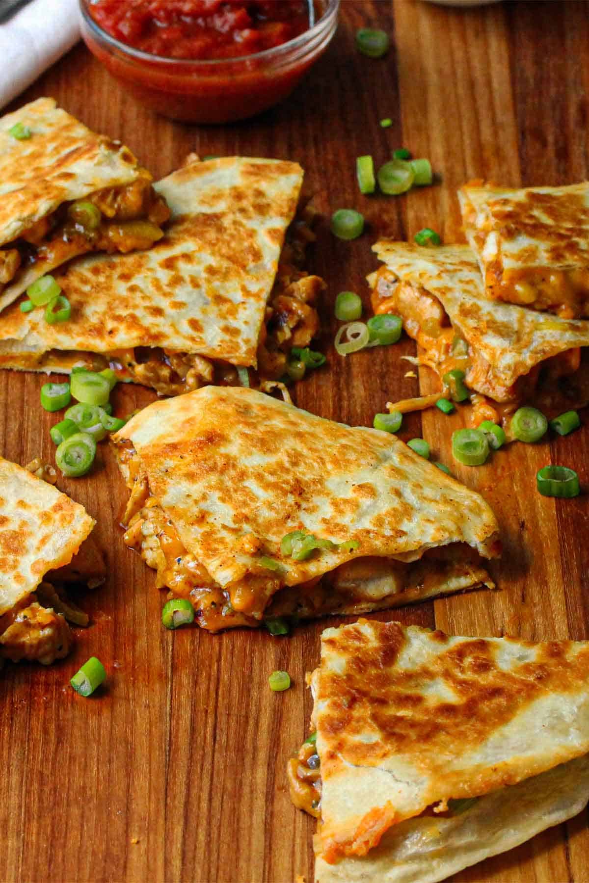 Triangle shapes of chicken quesadilla haphazardly scattered around on a large wooden cutting board.