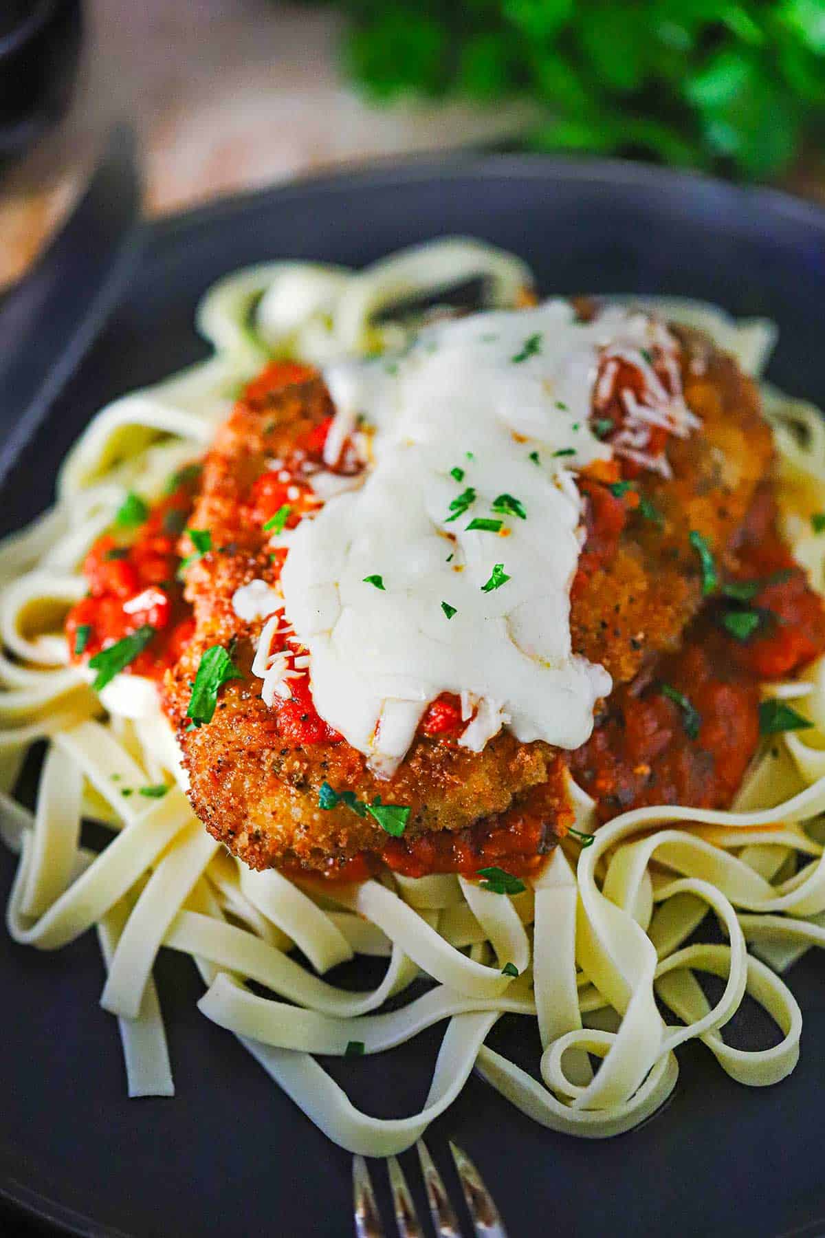 A black dinner plate filled with a serving of chicken Parmesan (Parmigiana) resting on a bed of cooked linguine and marinara sauce.