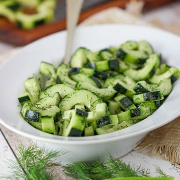 A white oval serving dish filled with sliced cucumber and dill salad with sprigs of fresh dill in front of the bowl.