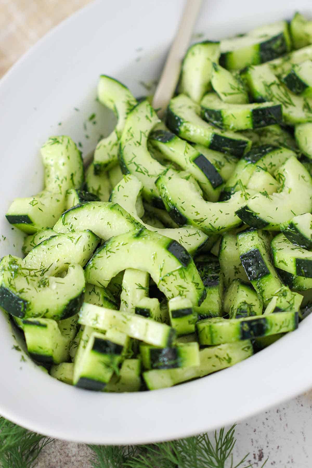 An white oval serving bowl filled with cucumber and dill salad with a spoon inserted on the side.