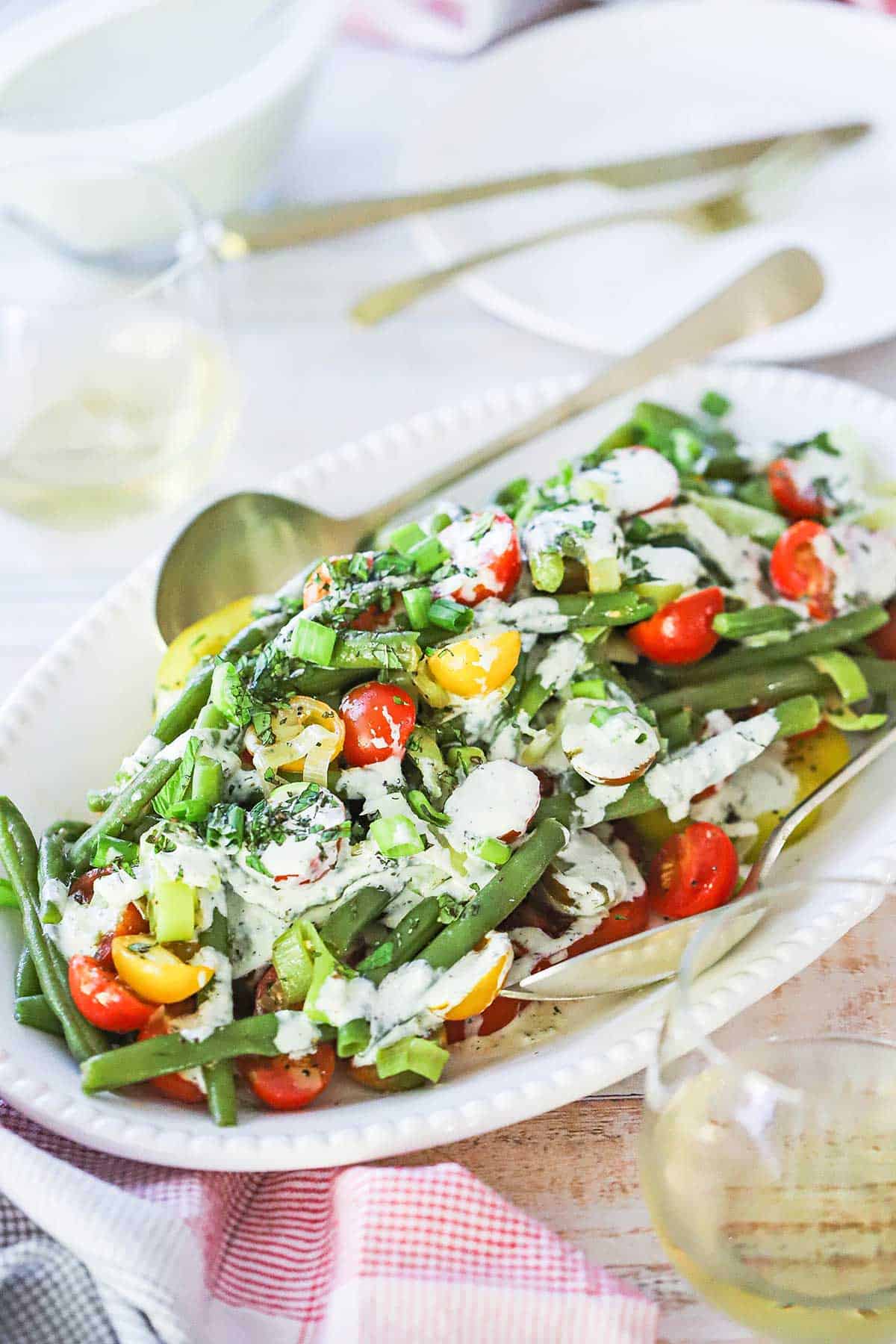An oval white platter filled with a large serving of green bean and tomato salad topped with a scallion buttermilk dressing, scallions, and chopped fresh herbs.