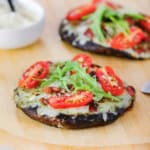 Two portobello BLO pizzas resting on a wooden pizza peel with both pizzas topped with melty Fontina cheese, sliced cherry tomatoes, and arugula leaves.