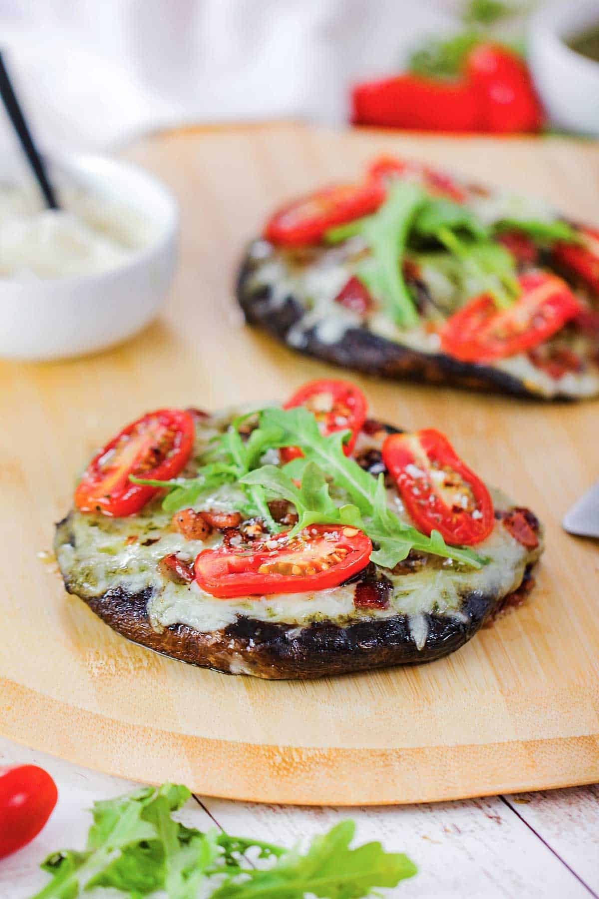 Two portobello BLO pizzas resting on a wooden pizza peel with both pizzas topped with melty Fontina cheese, sliced cherry tomatoes, and arugula leaves.