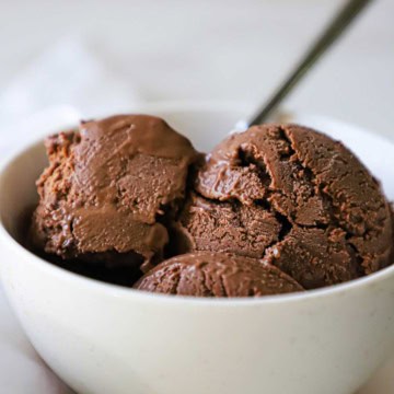 A white ice cream bowl filled with three scoops of homemade chocolate ice cream with a silver spoon inserted off to the side.