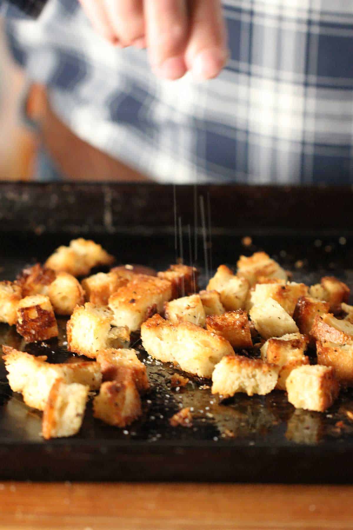 A person sprinkling coarse Kosher salt onto freshly baked homemade croutons that are resting on a black baking pan.