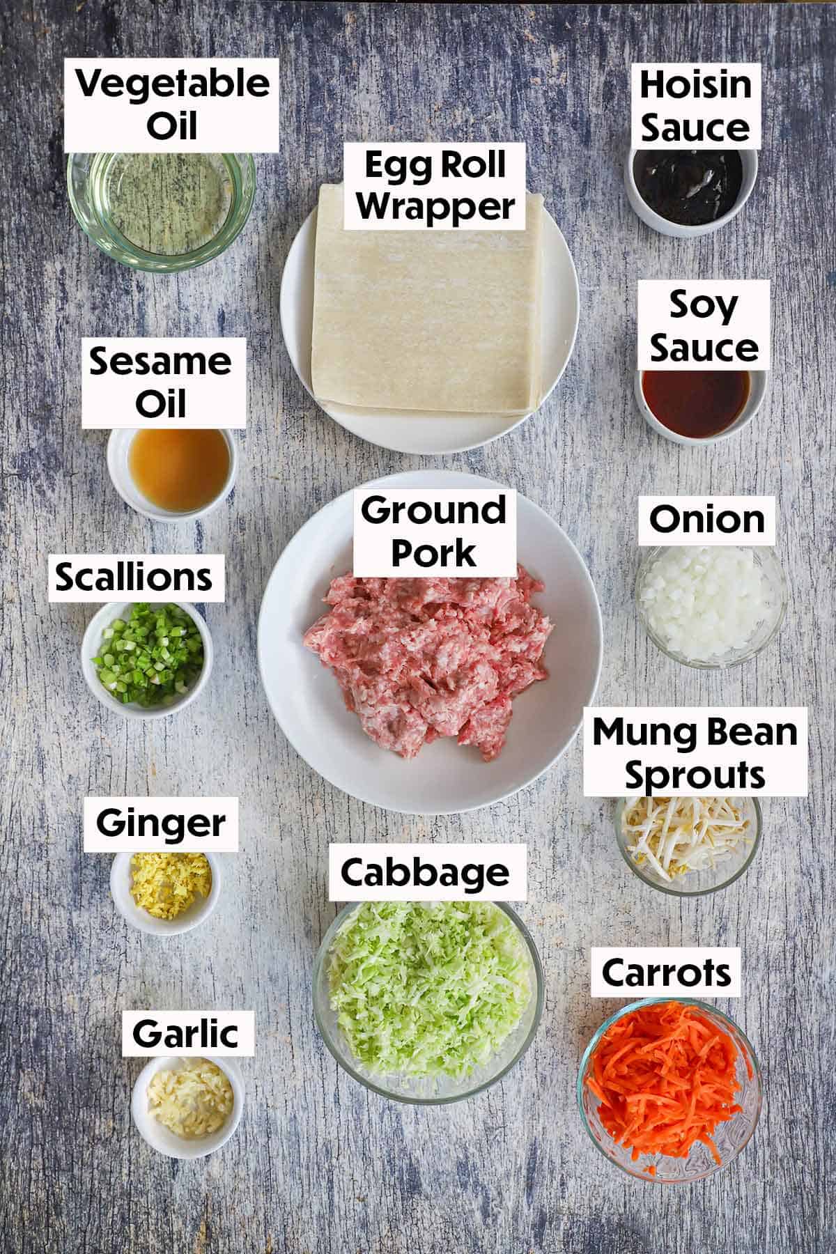 An arrangement of ingredients on a wooden grey background including ground pork, egg roll wrappers, cabbage, carrots, onion, garlic, ginger, and Asian seasonings.