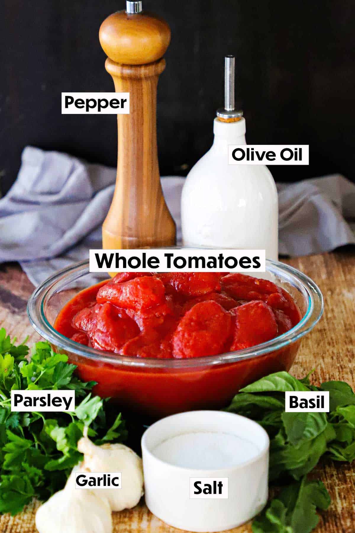 An arrangement of ingredients for homemade marinara sauce on a wooden board including a bowl of whole tomatoes, a peppercorn grinder, an olive oil dispenser, fresh Italian parsley, heads of garlic, fresh basil, and a container of salt.