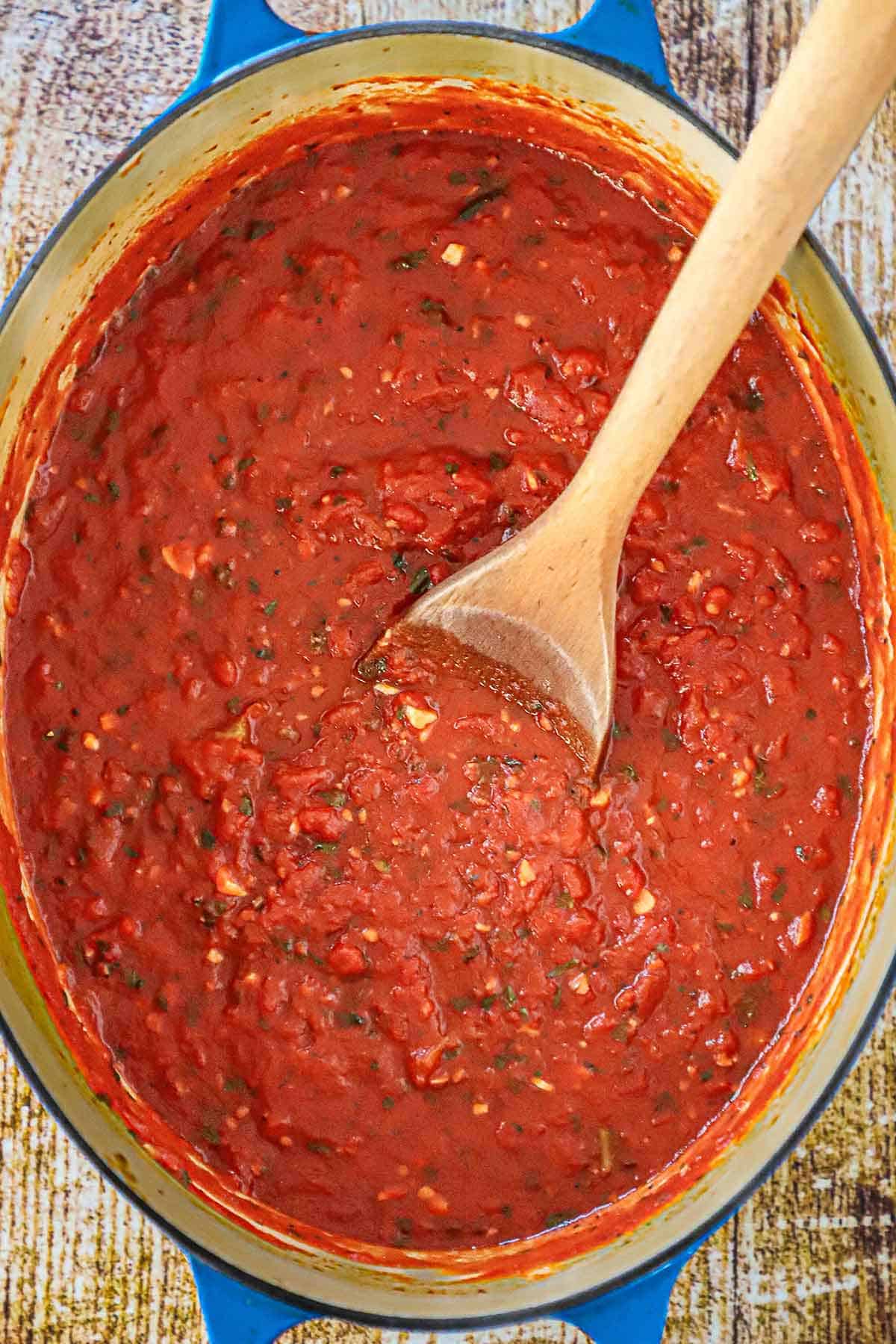 An overhead view of a large oval Dutch oven holding a freshly prepared batch of homemade marinara sauce with a large wooden spoon resting in the middle of the sauce with the handle resting on the side of the pot.