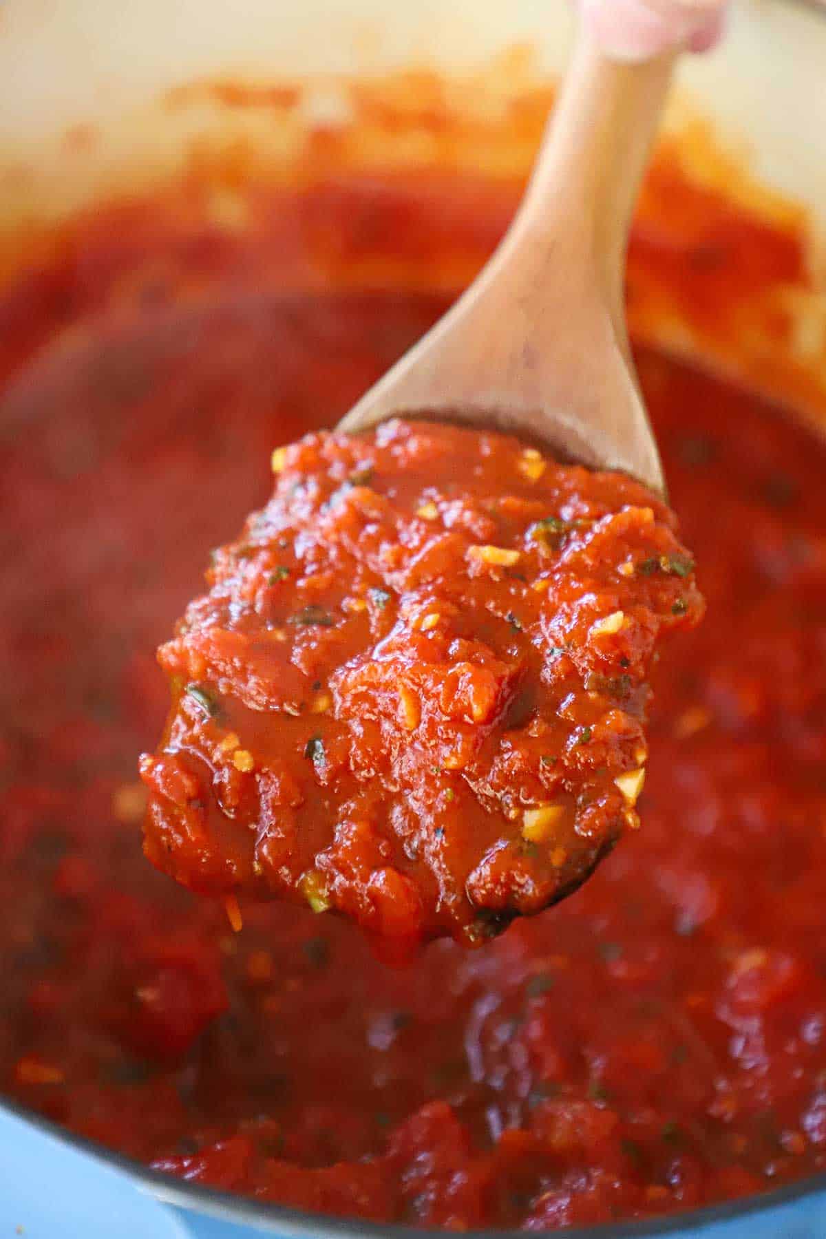 A wooden spoon holding up a large spoonful of homemade marinara sauce over a pot filled with the simmering sauce.