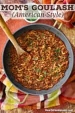 A deep skillet filled with Mom's goulash (American-style) with a wooden spoon inserted into it with a small bowl of chopped parsley sitting nearby.