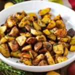 A white serving bowl filled with crispy roasted potatoes with balsamic and herbs with stems of fresh herbs nearby.