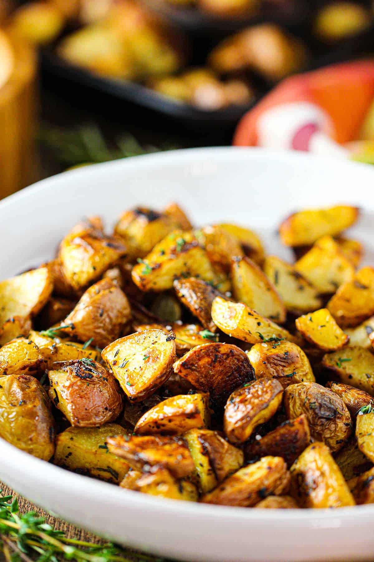 A white serving bowl filled with crispy roasted potatoes with balsamic and herbs with stems of fresh herbs nearby.