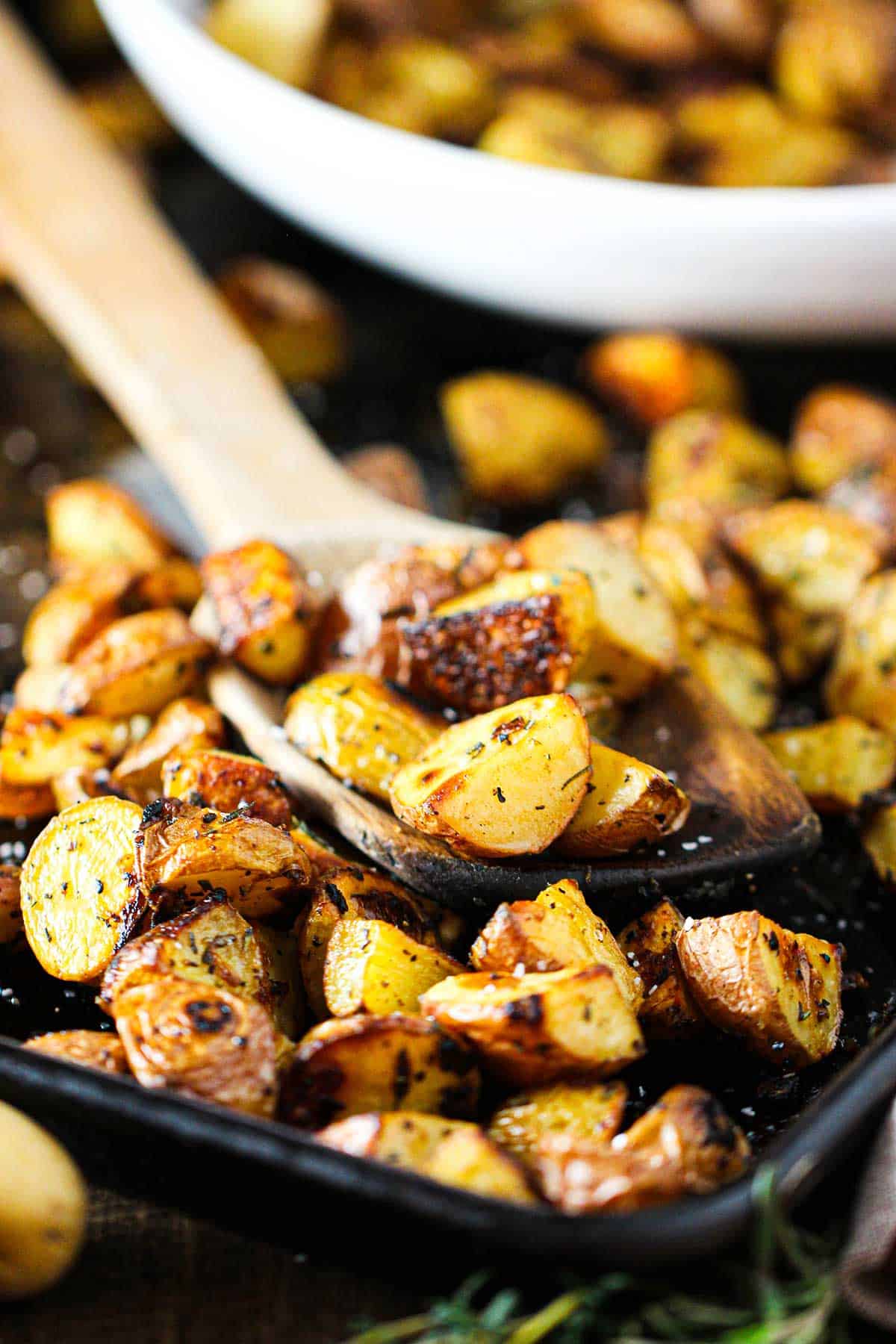 Crispy roasted potatoes with balsamic and herbs in a black baking sheet pan with a wooden spoon holding some of the potatoes in the middle of the pan.