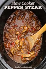 An overhead view of the inside of a slow cooker that is filled with pepper steak in a dark brown gravy with a wooden spoon in the middle of the sauce.